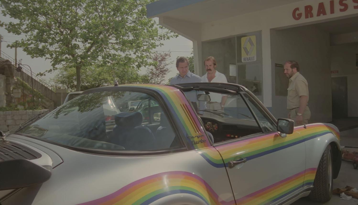 Ein silbernes Auto mit Regenbogenstreifen steht in der Nähe einer Garage. Drei Männer stehen daneben. Ein Renault-Schild ist sichtbar.