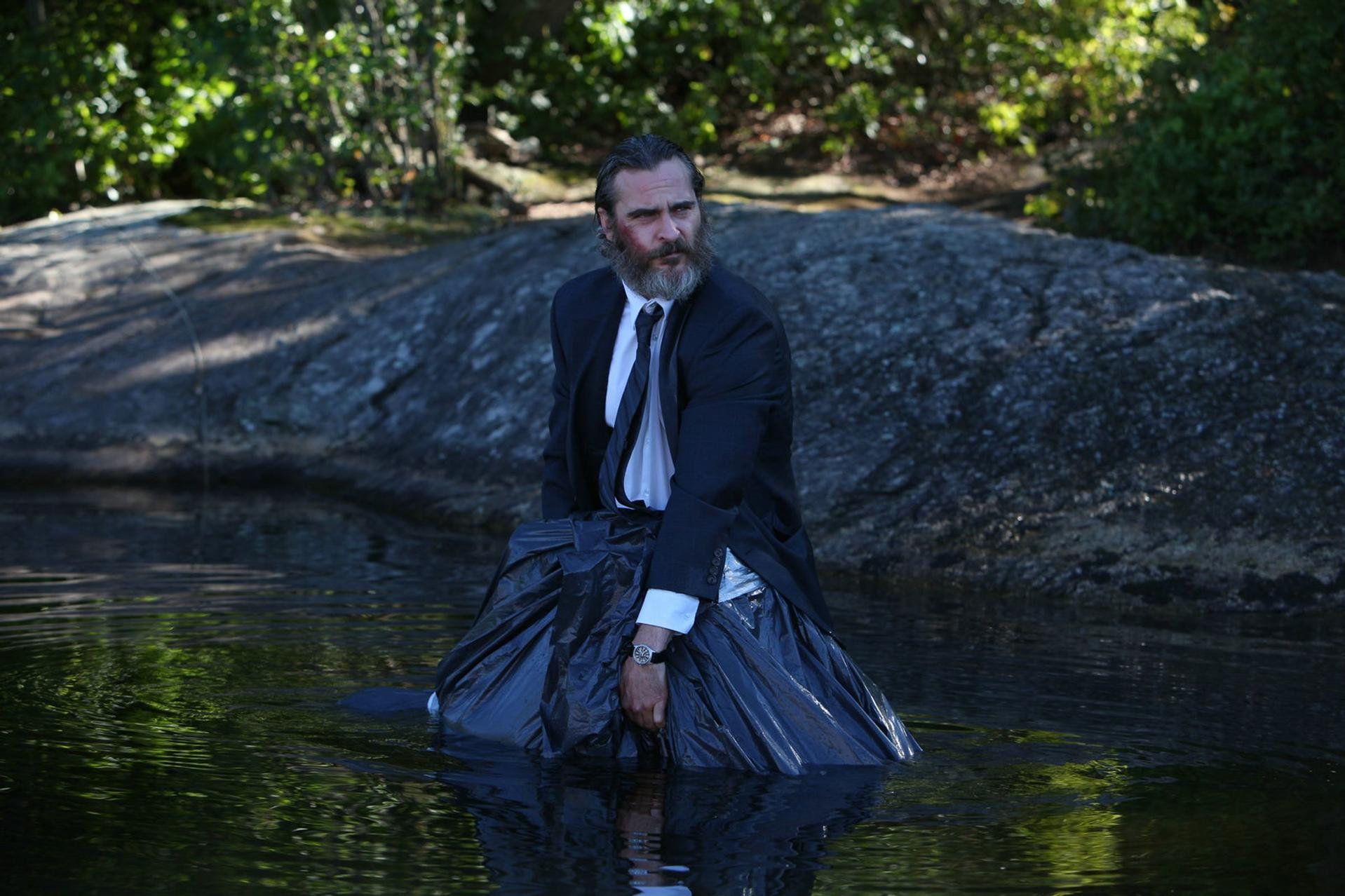 Ein Mann im Anzug, eingehüllt in einen schwarzen Müllsack, sitzt im Wasser. Bäume und ein großer Felsen im Hintergrund.