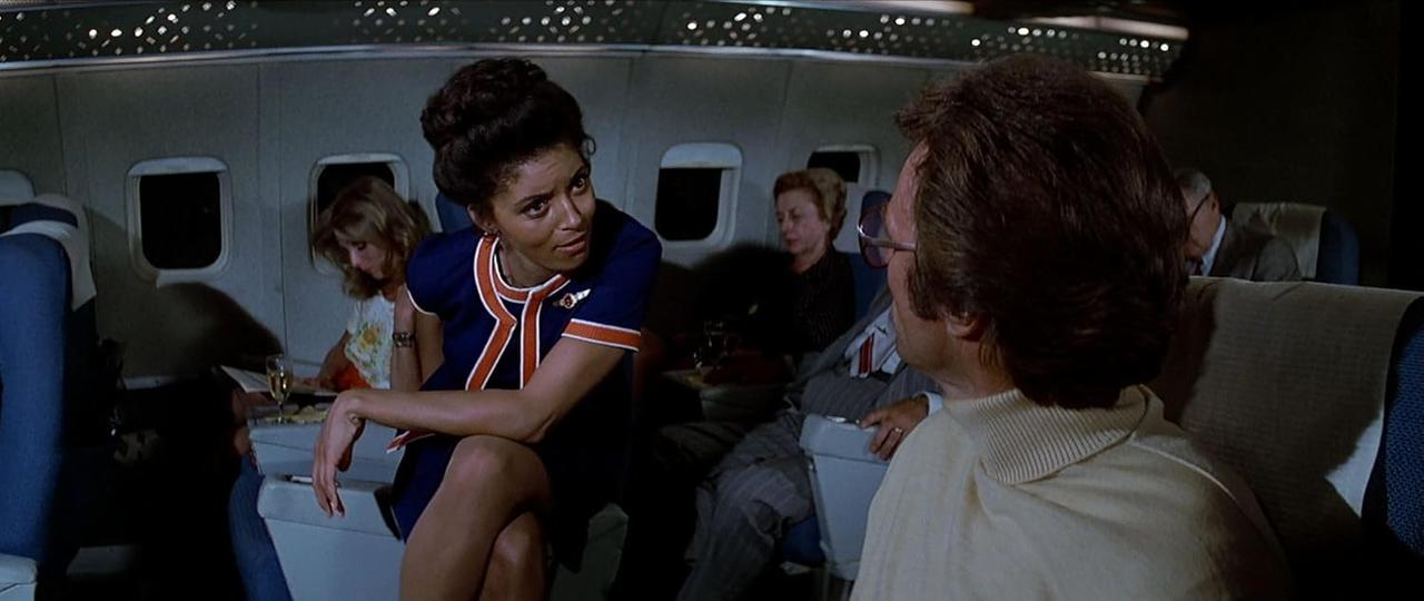 Eine Stewardess in blauer Uniform spricht mit einem Mann in einem Flugzeug. Passagiere sitzen hinter ihnen.