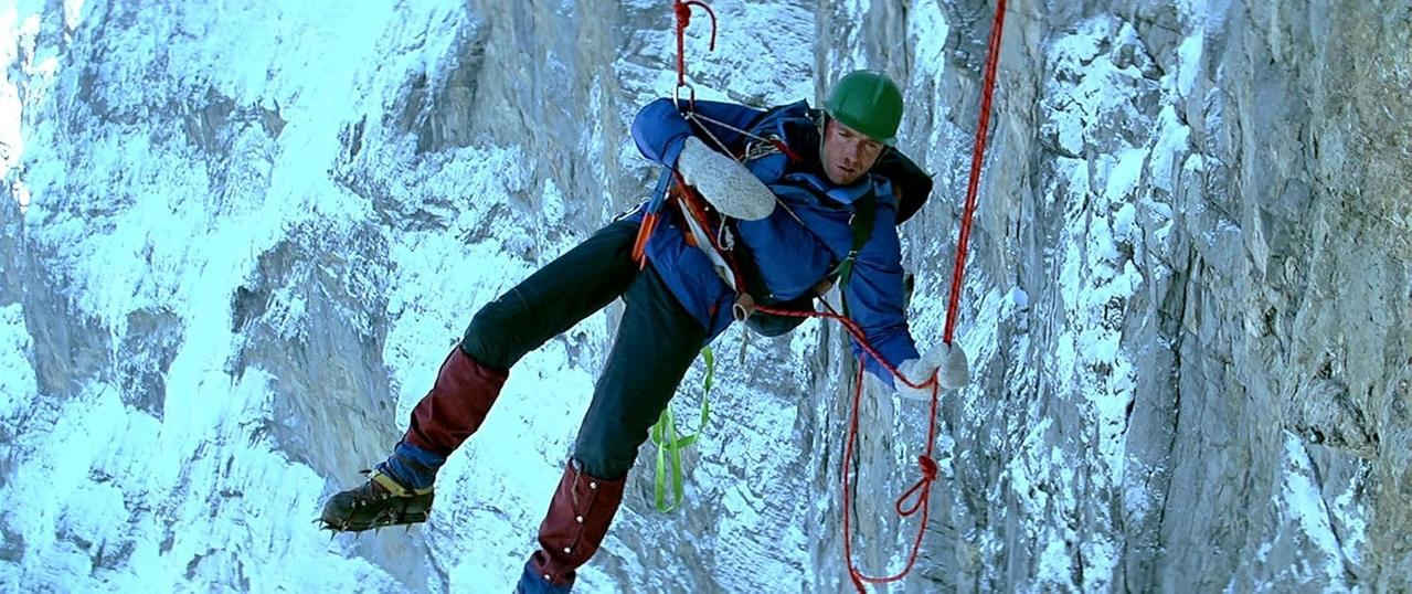 Mann seilt sich an einer Felswand ab. Er trägt einen Helm, einen blauen Anzug und Kletterschuhe. Rote Seile sichern ihn.