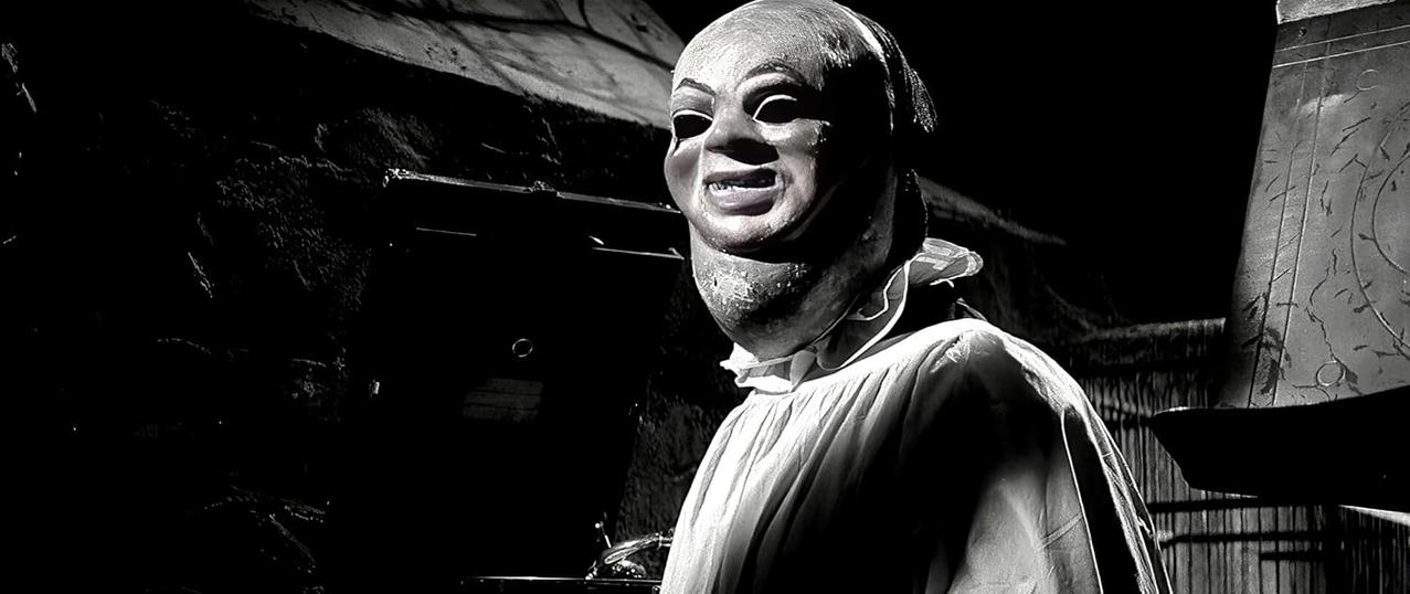Eine Person mit Maske, weitem Hals und weißem Hemd. Der Hintergrund ist dunkel, mit einer Wand.