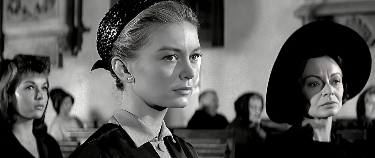 Schwarz-Weiß-Bild von Frauen in einer Kirche. Eine Frau mit Hut, eine andere mit Kopftuch. Andere Figuren sind im Hintergrund verschwommen.