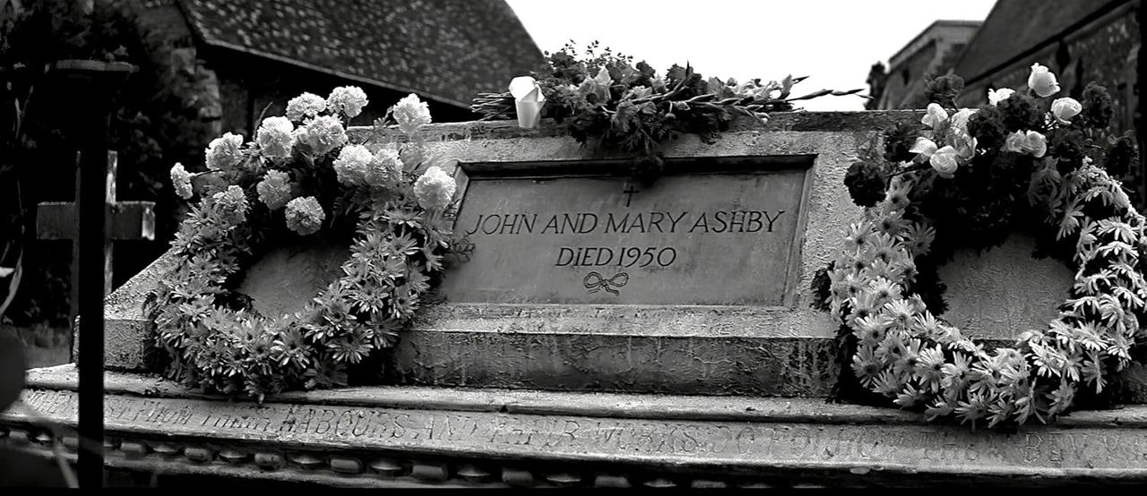 Graustufenbild eines Grabsteins mit der Inschrift "John and Mary Ashby Died 1950", umgeben von Blumengirlanden.