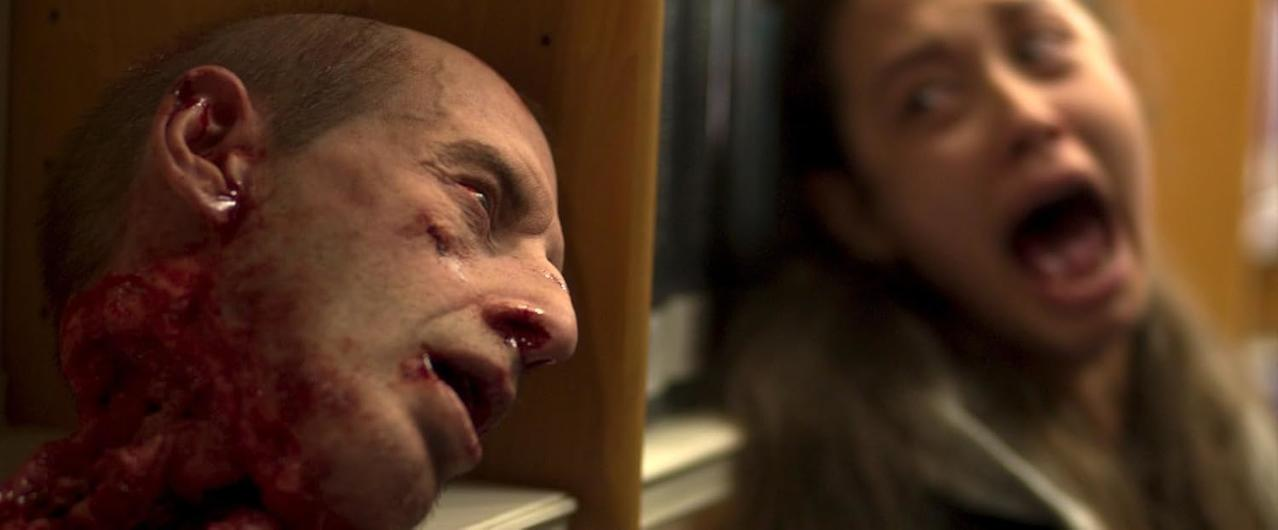 Ein abgetrennter Kopf mit Blut. Eine Frau schreit im Hintergrund. Die Szene deutet auf Gewalt und Horror hin.