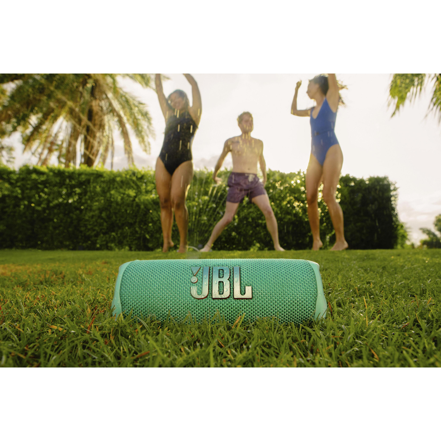 Three people jump in front of a green JBL speaker on grass. They are wet, and a sprinkler is in the background. 
