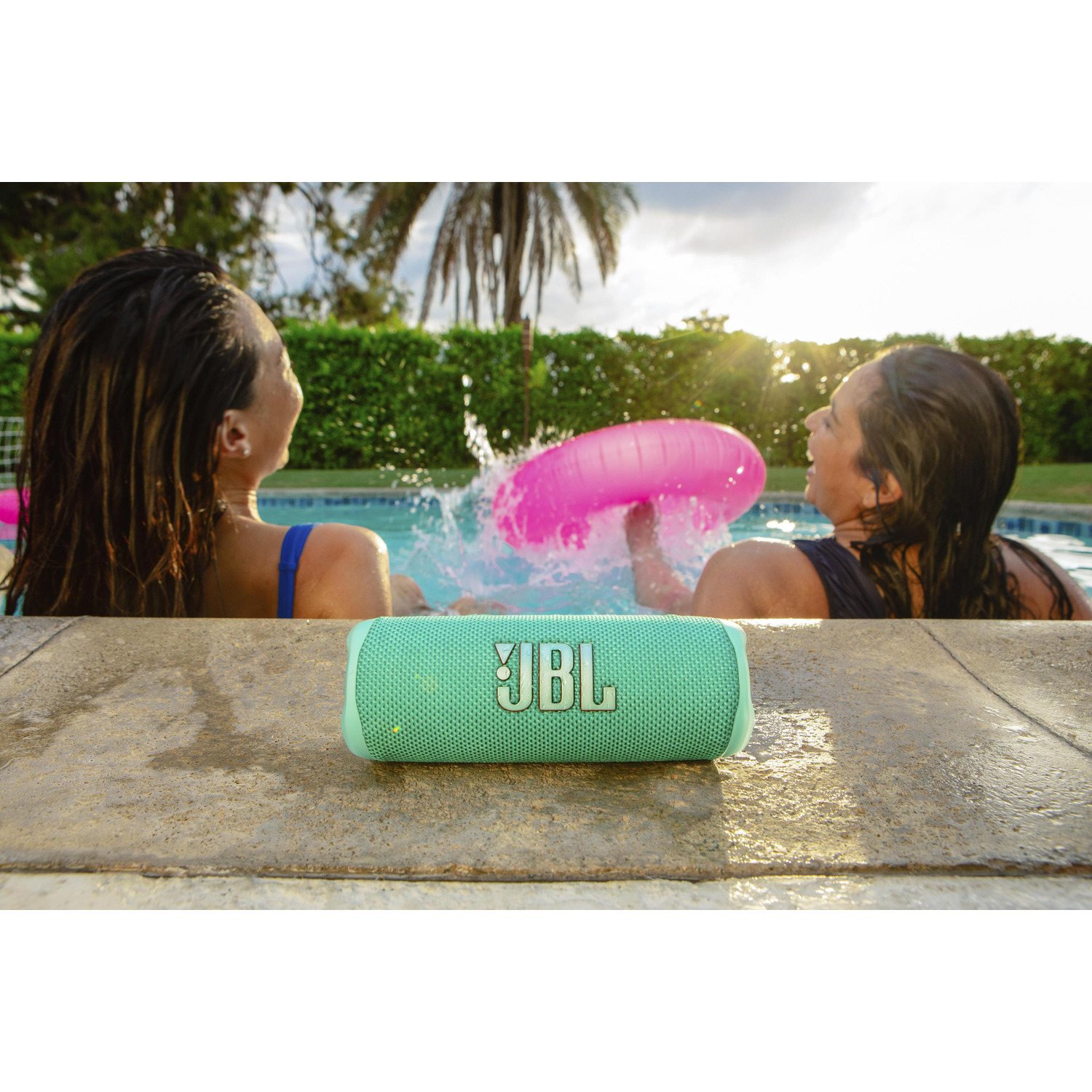 Two women in a pool, one with a pink floatie. A JBL speaker sits on the edge. Water splashes. Green trees and sun in background.