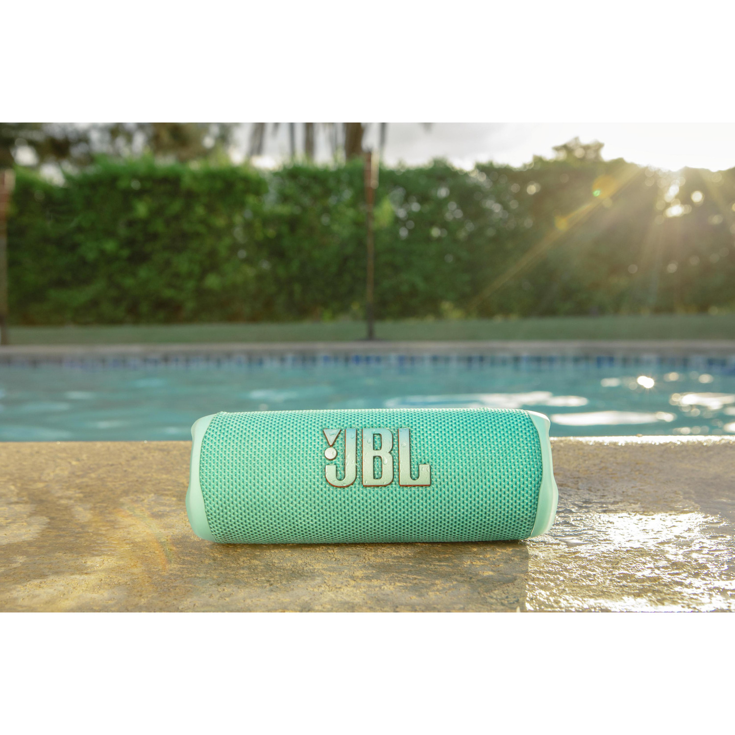 A JBL speaker rests poolside. The teal speaker is on a concrete ledge with the pool and green foliage in the background.