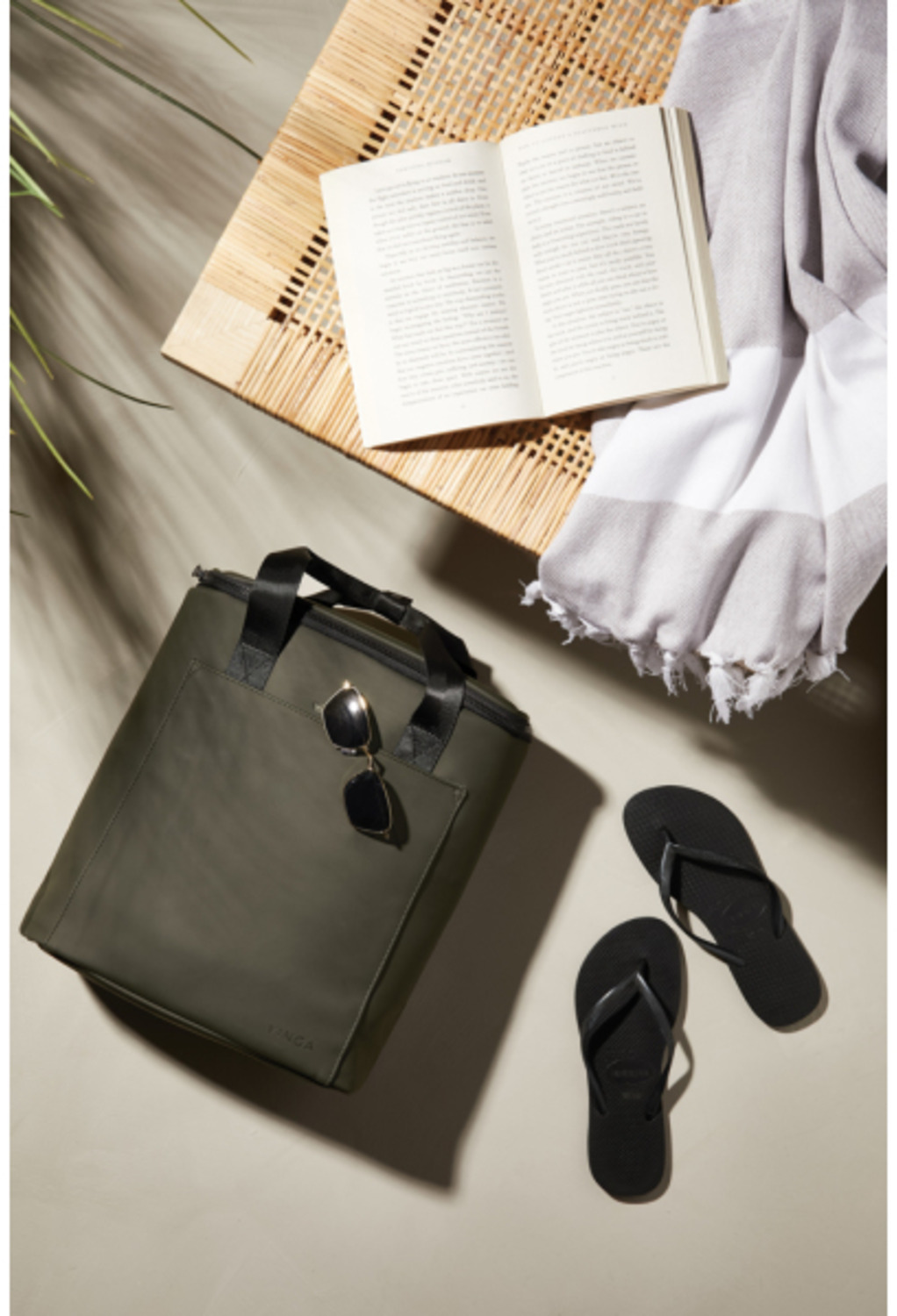 A beach scene with a tote bag, sunglasses, flip-flops, a book, and a towel on a woven table.