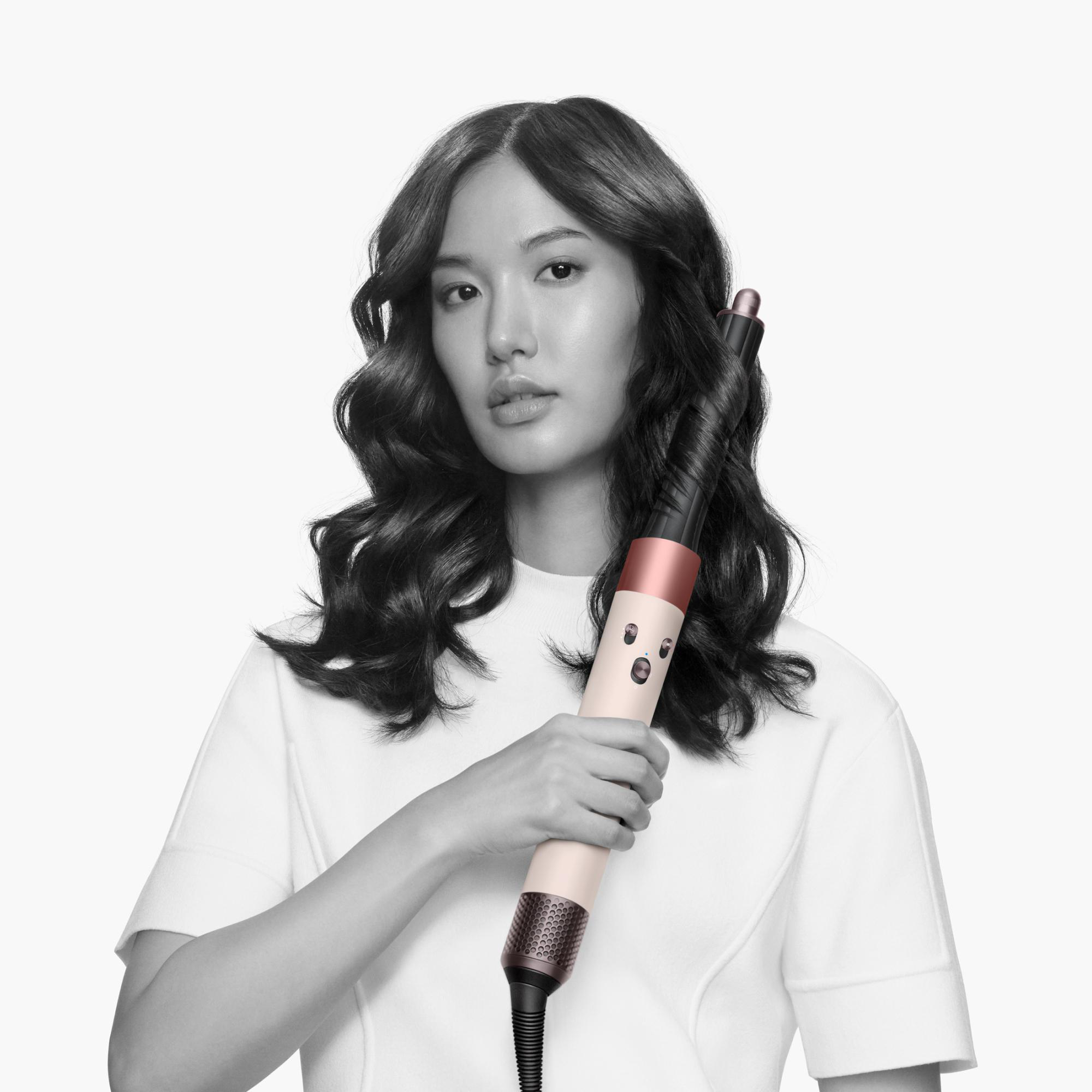 Eine Frau mit dunklem Haar lockt ihr Haar mit einem Styler. Sie trägt ein weißes Oberteil. Der Hintergrund ist weiß.