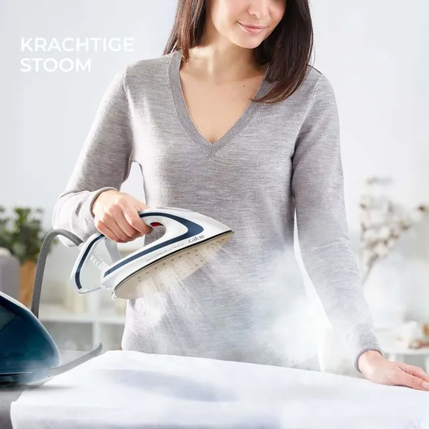 A woman ironing a white cloth with a steam iron. The iron is white and blue, steam is visible. The woman is wearing a grey sweater.