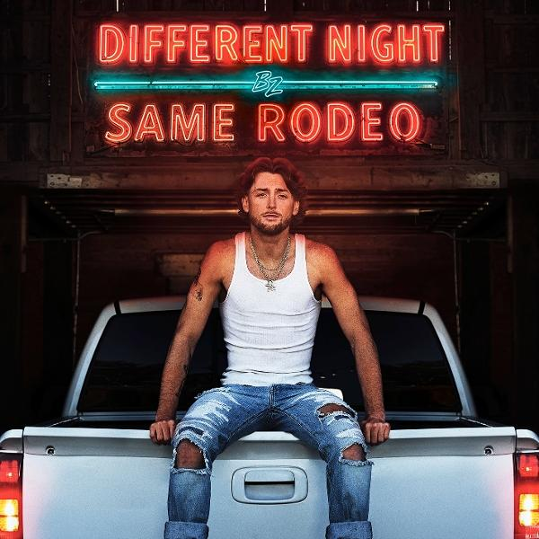 Egy férfi egy teherautón ül, a 'Different Night, Same Rodeo' feliratú neonfény alatt. Fehér trikót és szaggatott farmert visel.