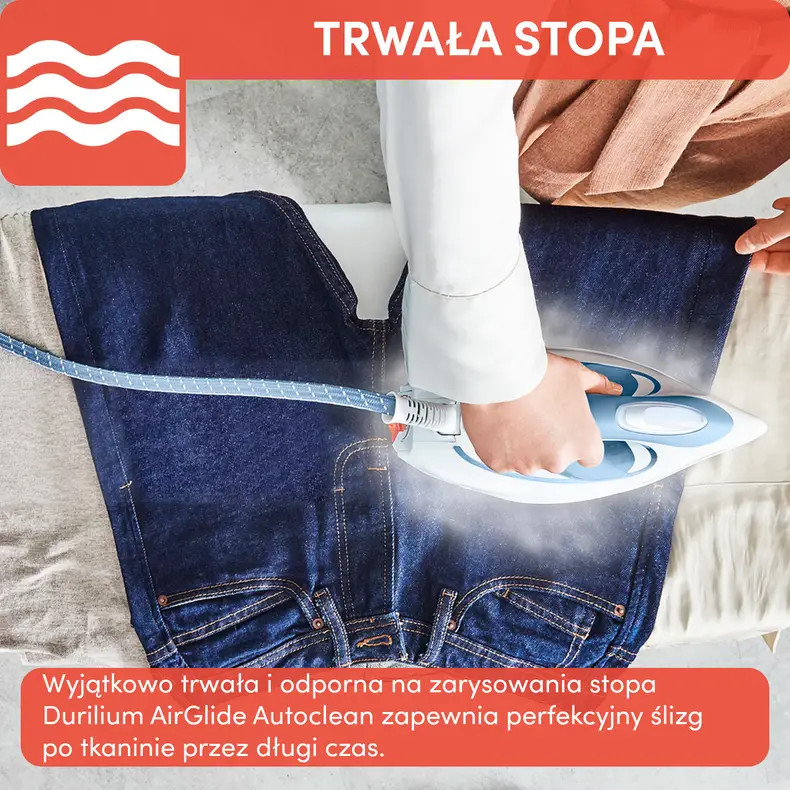 Osoba prasuje ciemnoniebieskie dżinsy białym i niebieskim żelazkiem. Para unosi się, a żelazko znajduje się na desce do prasowania.