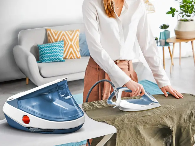 Kobieta prasuje koszulę na desce do prasowania. Żelazko z generatorem pary stoi obok deski. W tle kanapa i dekoracje.