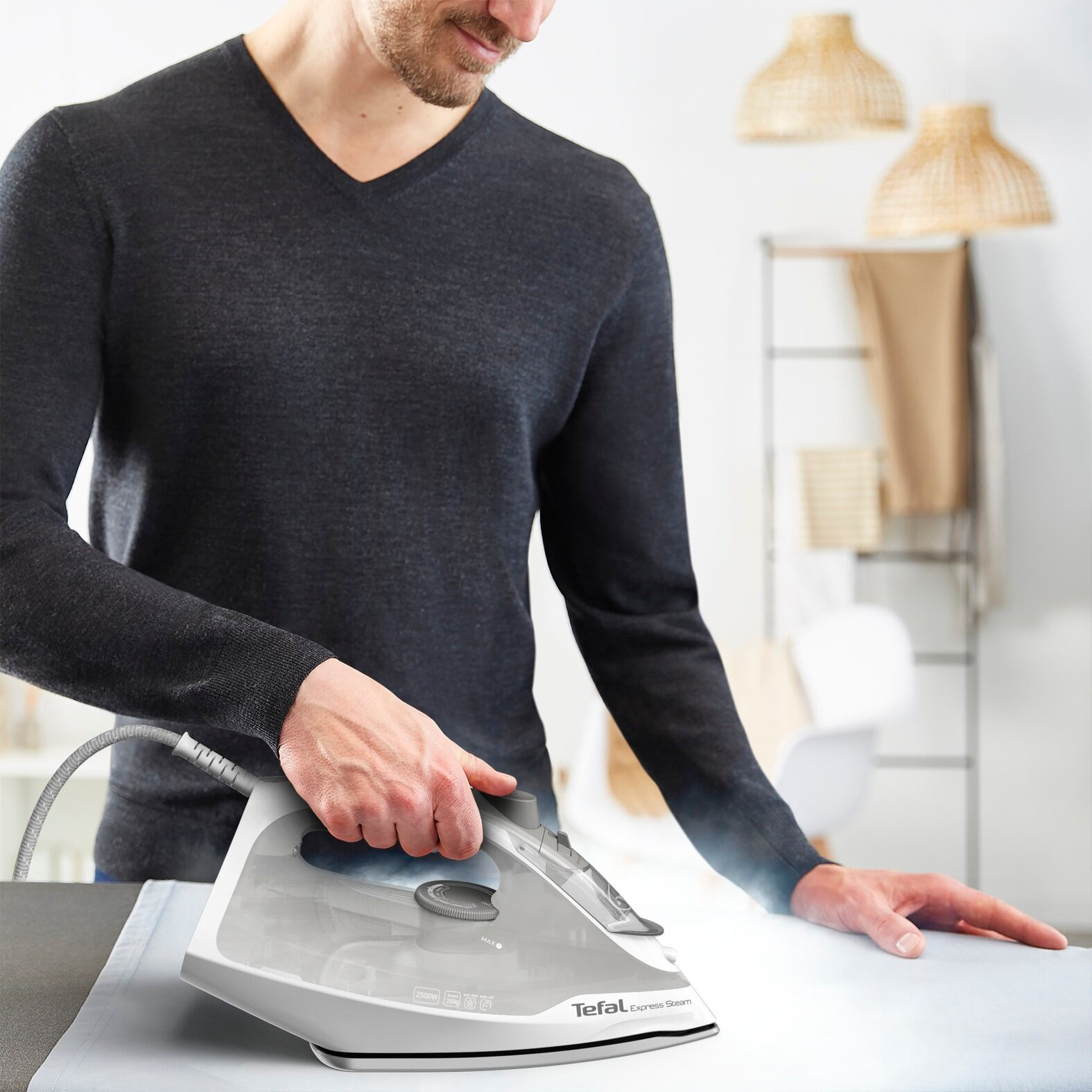 Mężczyzna prasujący żelazkiem parowym. Szary sweter, biały stół i jasne tło z lampami i ubraniami.