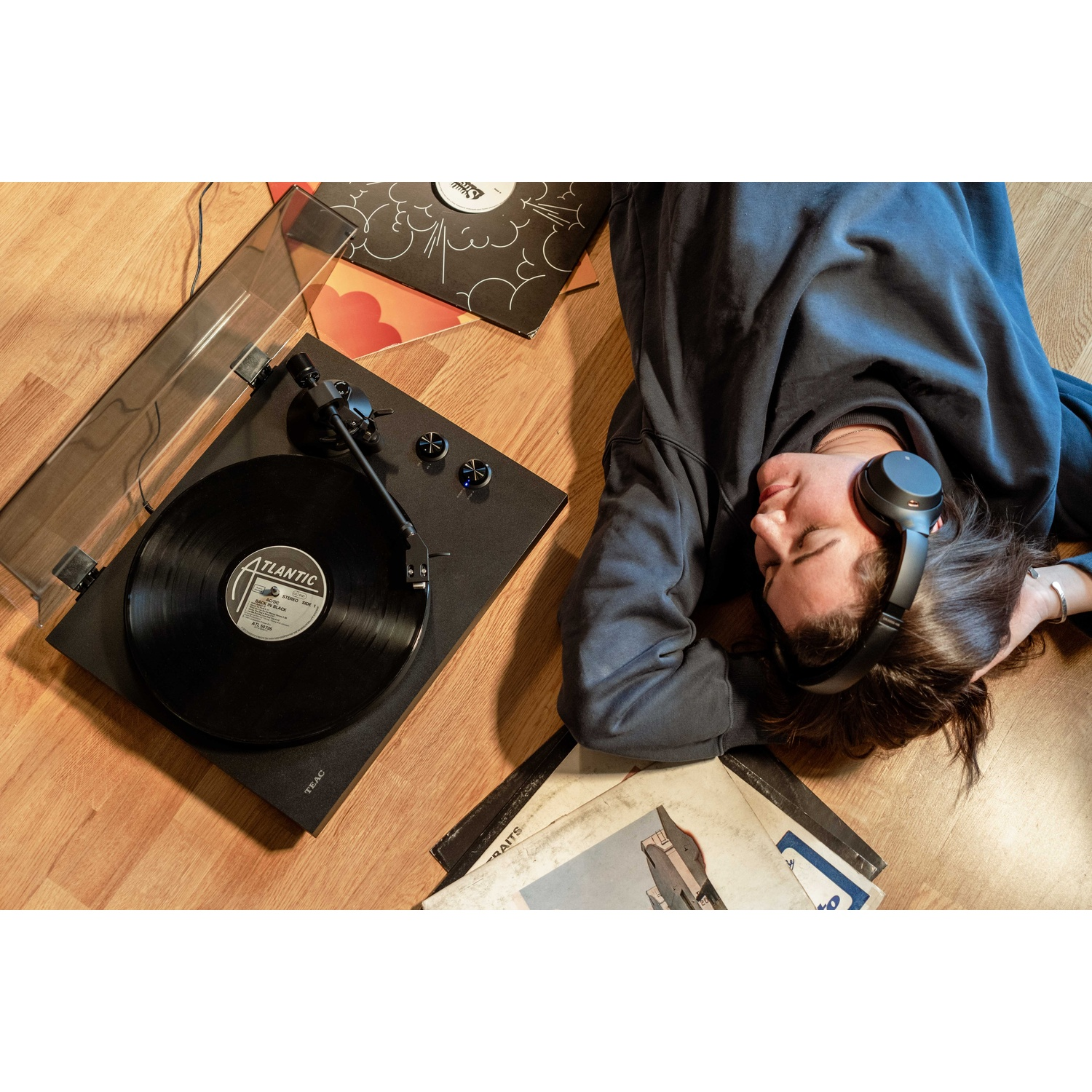 Una persona con le cuffie è sdraiata su un pavimento di legno vicino a un giradischi e dischi in vinile.