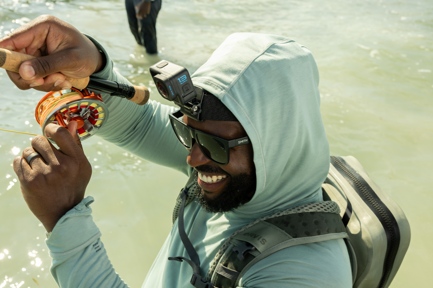 Un hombre con gafas de sol y una sudadera con capucha está pescando. Sonríe mientras sostiene una caña de pescar y un carrete. Una cámara está montada en su cabeza.