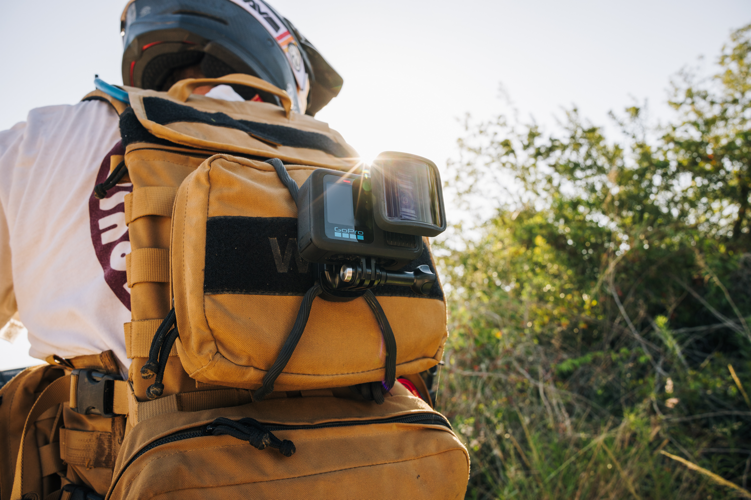 Una persona usa un casco y una mochila con una cámara GoPro adjunta. La mochila es de color canela y el fondo es soleado con follaje verde.