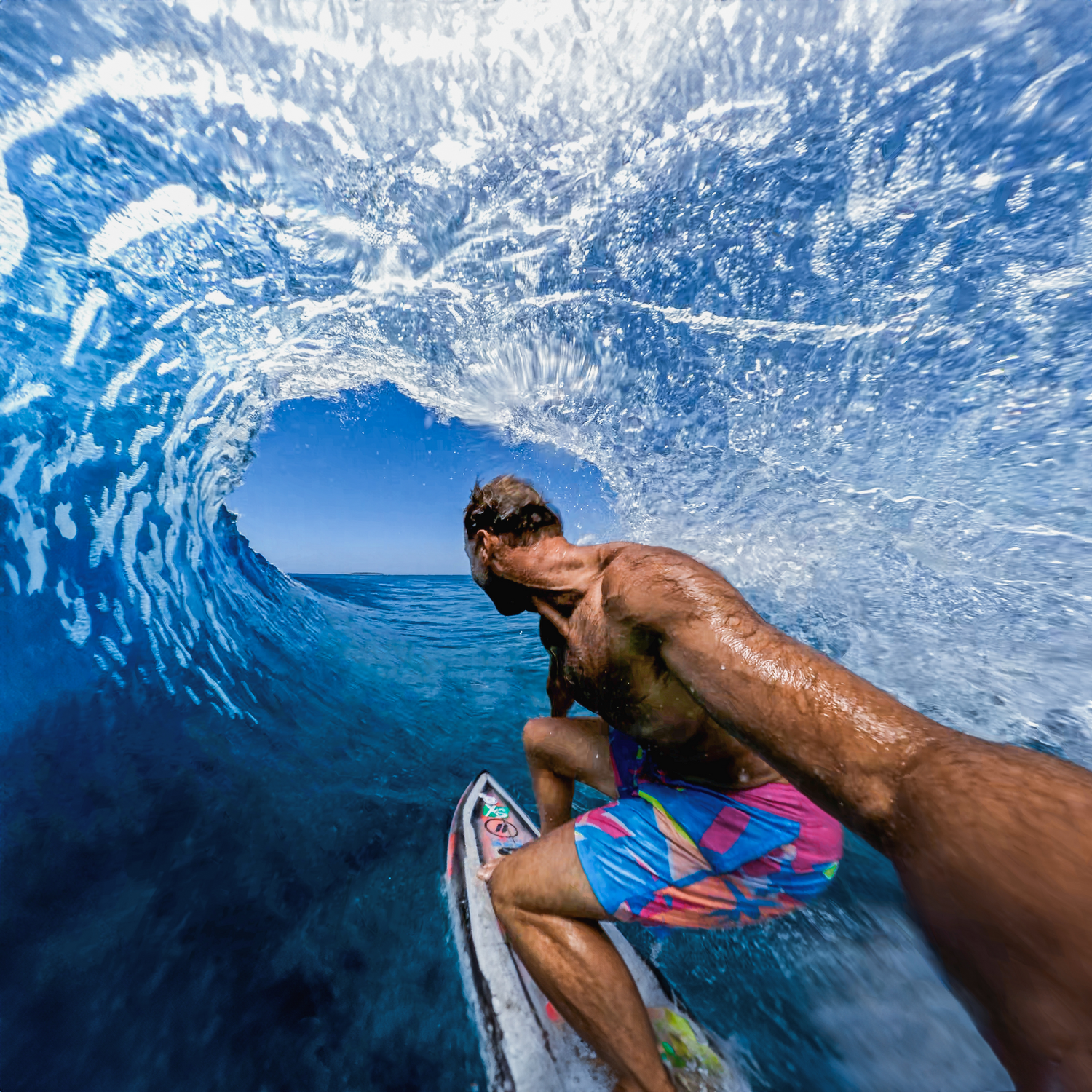 Un surfista surfea una ola. La ola es un enorme tubo azul. El surfista está en una tabla, usa pantalones cortos coloridos y tiene piel morena.