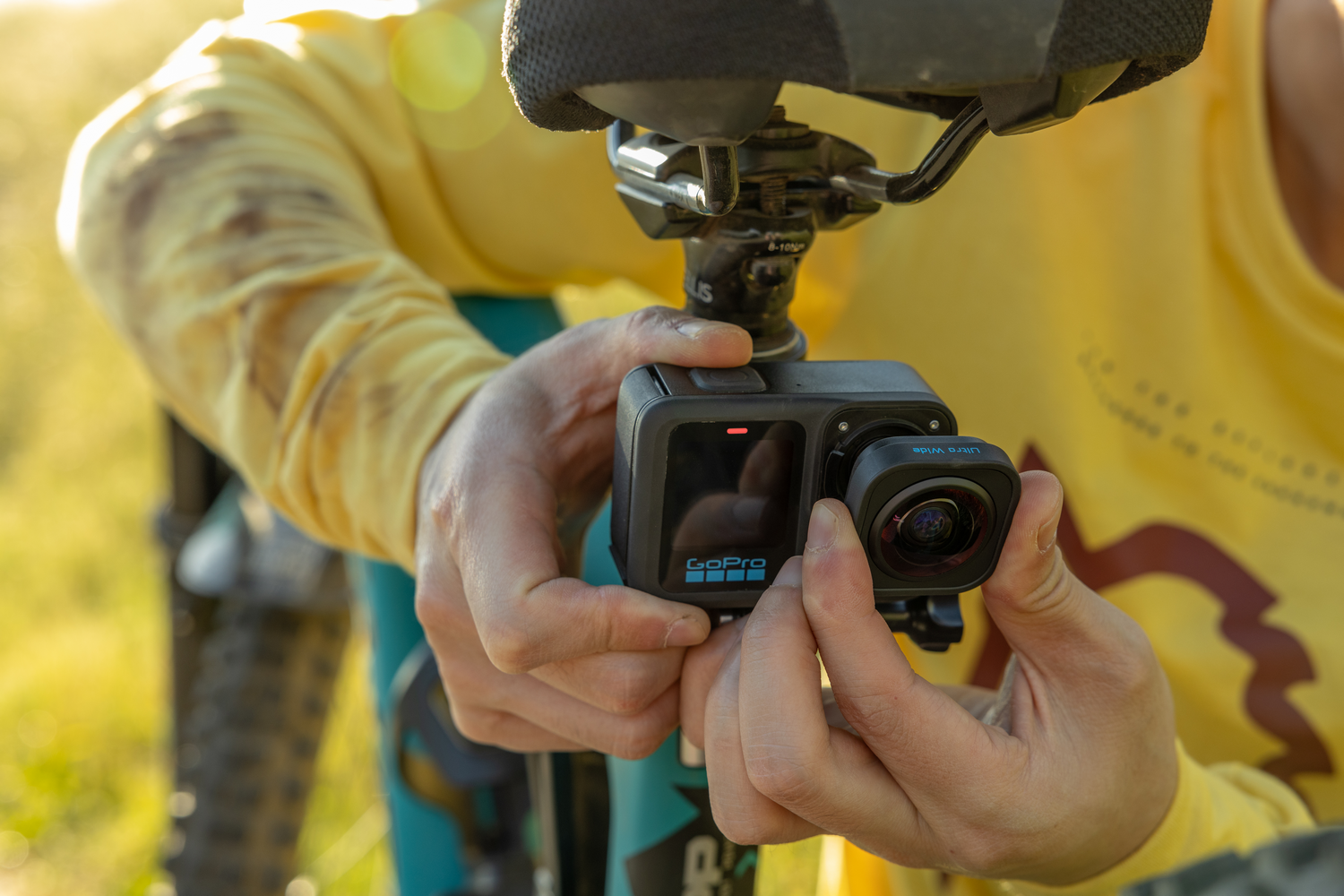 Una persona sostiene una cámara GoPro sujeta al asiento de una bicicleta. La cámara es negra y la persona lleva una camisa amarilla de manga larga.