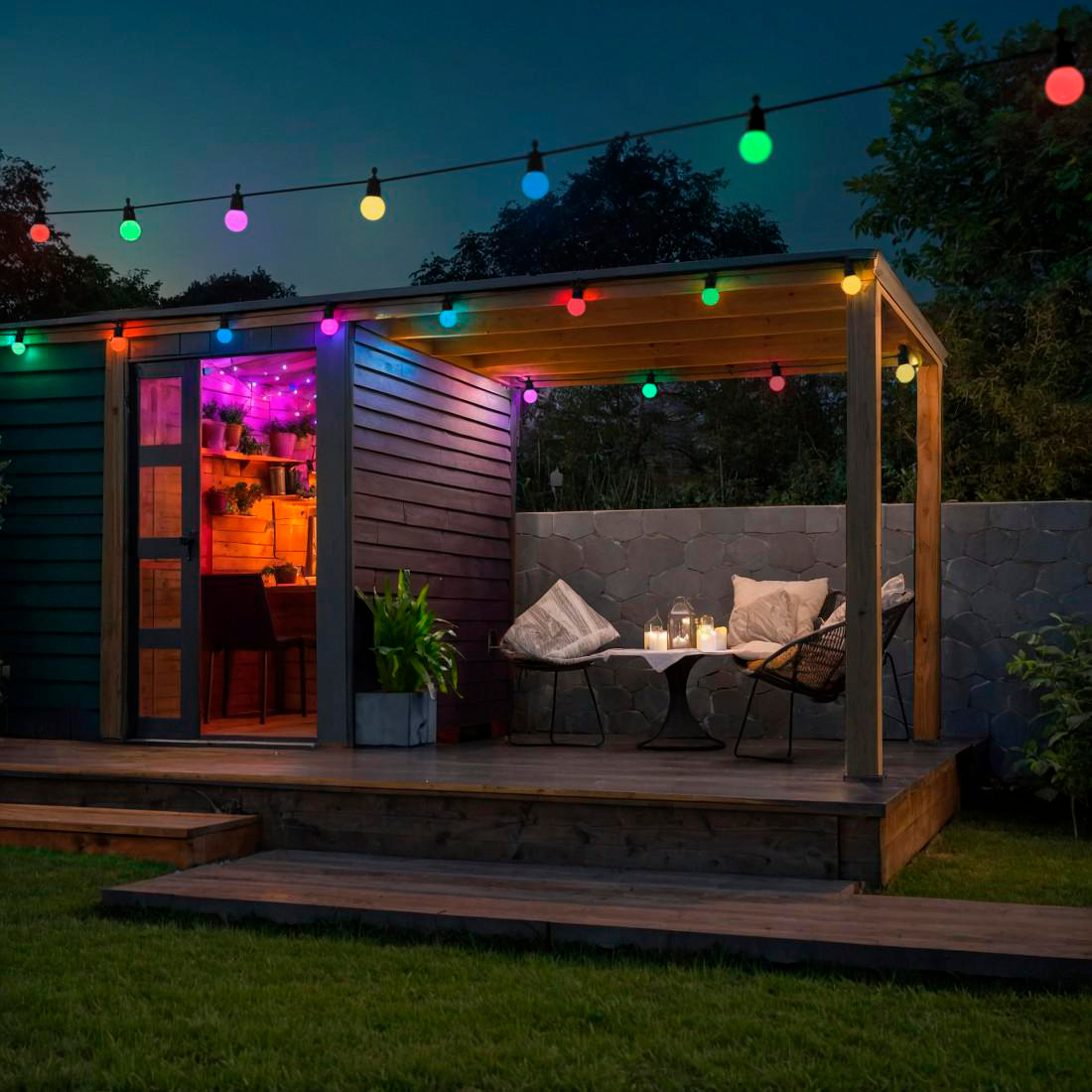 Außenszene bei Nacht. Schuppen mit offener Tür, bunte Lichter, Tisch, Stühle und Pflanzen. Grünes Gras im Vordergrund.