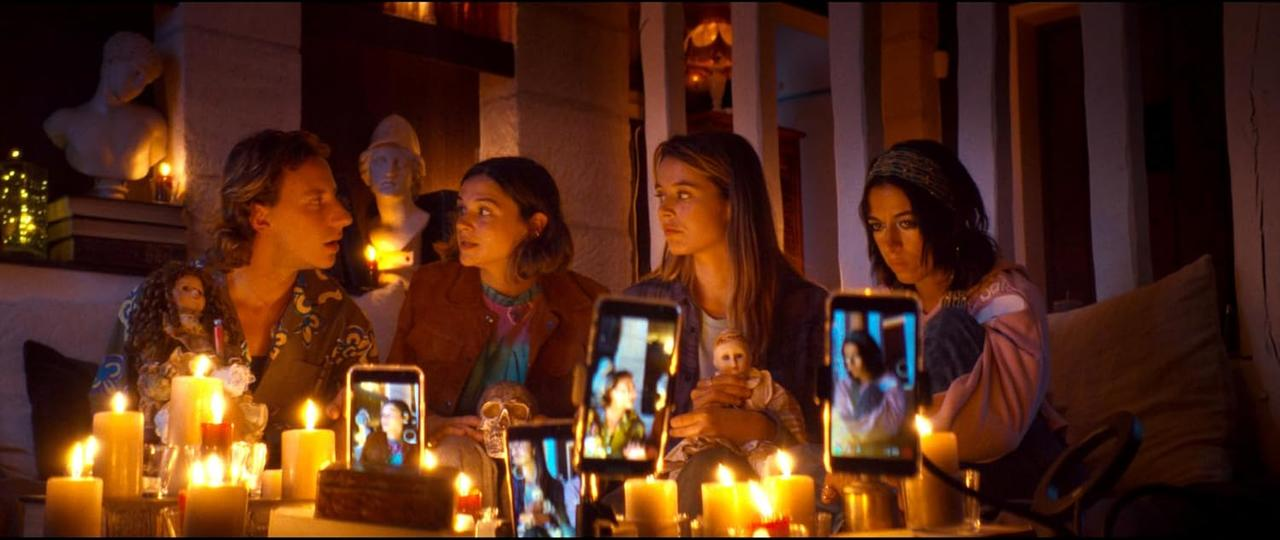 Vier Personen versammeln sich um brennende Kerzen und halten Smartphones. Statuen und ein Schädel sind sichtbar. Die Szene ist schwach beleuchtet.