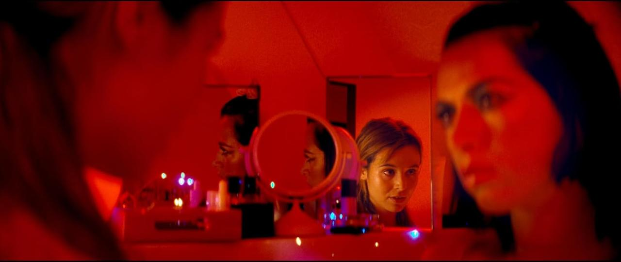 Eine Frau schaut in einen Spiegel, roter Raum. Eine andere Frau ist im Hintergrund zu sehen. Schminkartikel auf dem Tisch. Rotes Licht.