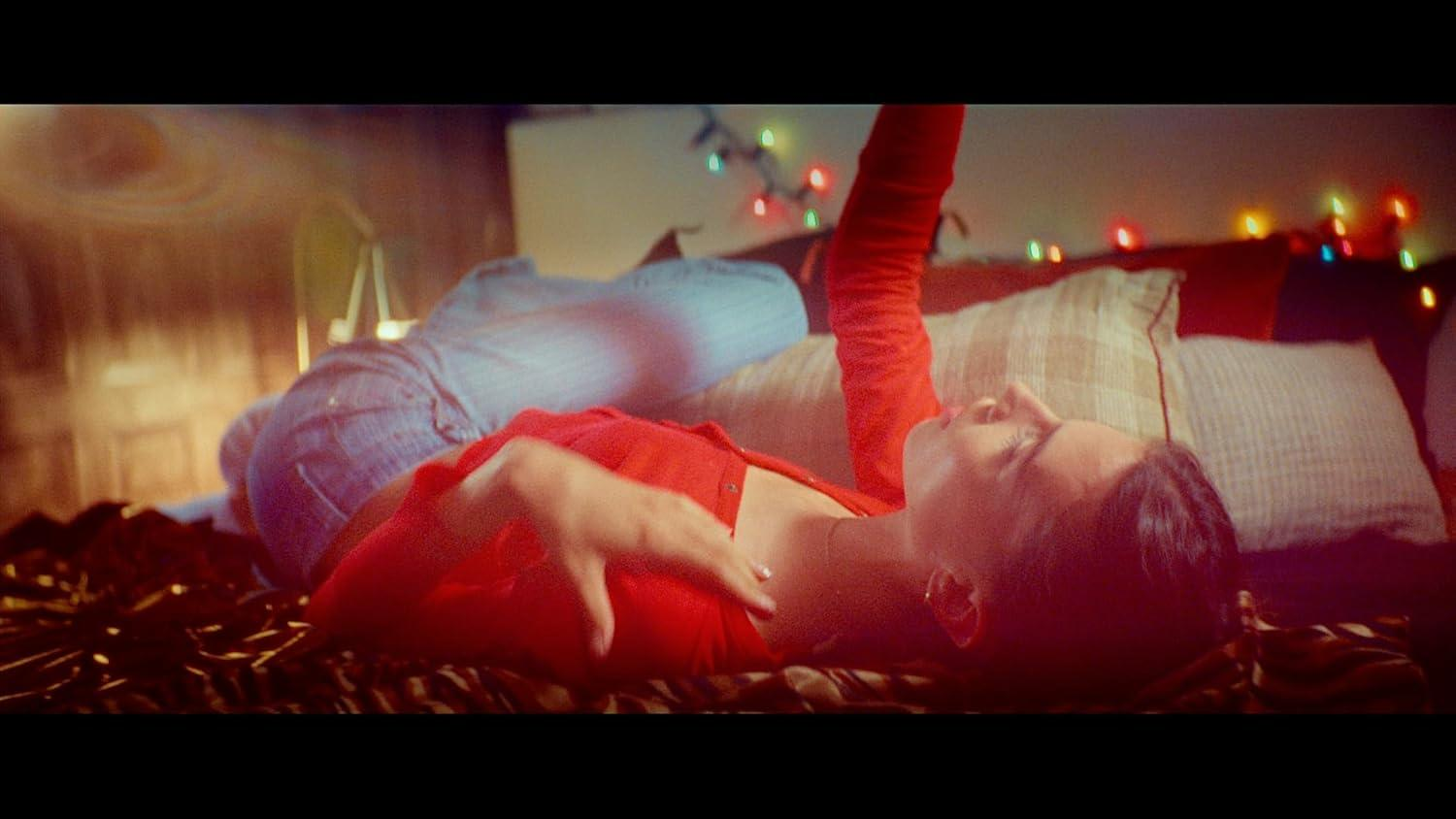 Eine Person liegt auf einem Bett und trägt Rot. Bunte Lichter sind im Hintergrund. Die Person ruht auf Kissen.