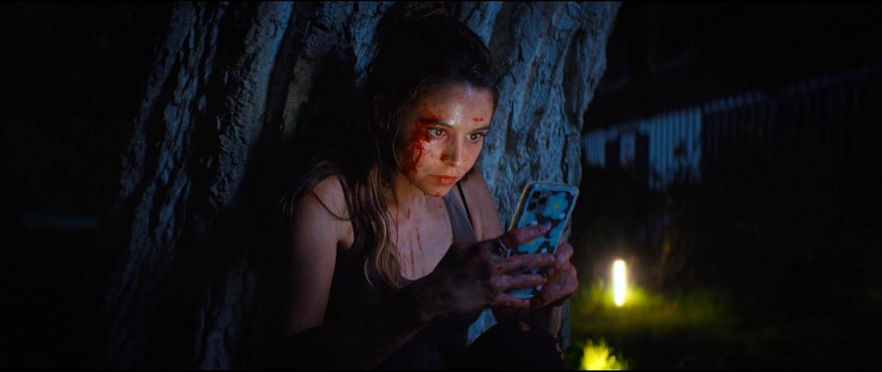 Eine Frau mit Blut im Gesicht blickt auf ein Telefon. Sie steht in dunkler Umgebung an einem Baum. Im Hintergrund ist ein Licht zu sehen.