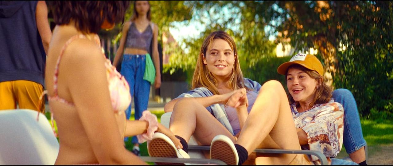 Mehrere junge Frauen sitzen im Freien. Eine trägt einen Hut. Andere tragen Freizeitkleidung. Eine Person im Bikini-Oberteil steht im Fokus.