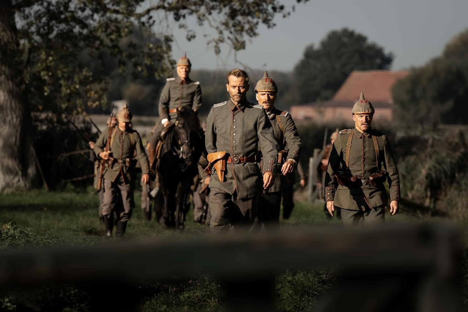 Soldaten in grauen Uniformen marschieren vorwärts auf einem Feld. Einige reiten auf Pferden. Einer hat ein Holster. Bäume und ein Gebäude sind im Hintergrund.
