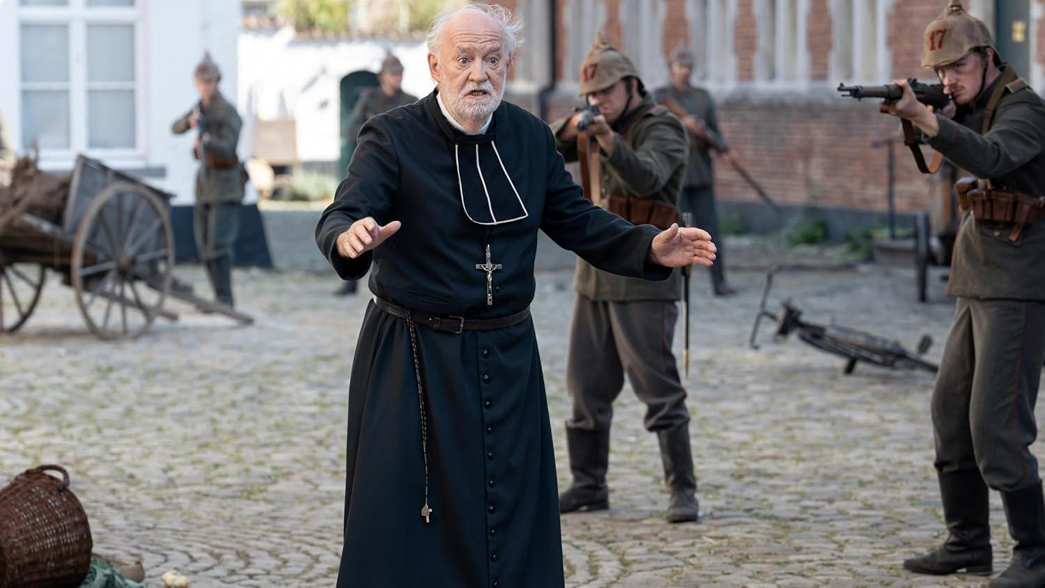 Ein Priester in schwarzen Gewändern gestikuliert, Soldaten in WK I-Uniformen zielen mit Gewehren. Ein Wagen und Kopfsteinpflaster im Vordergrund.