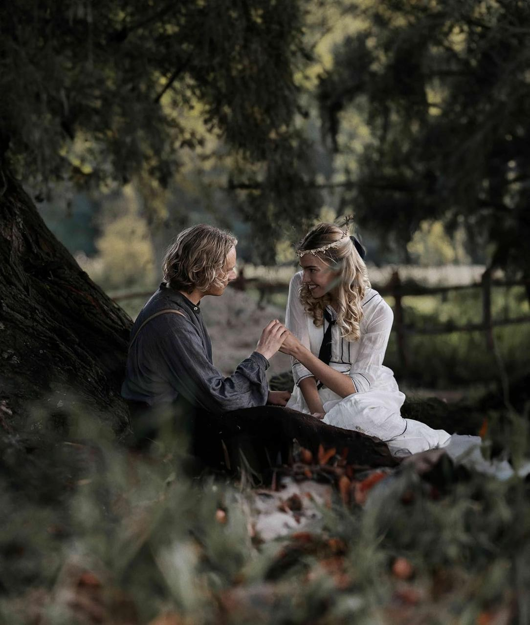 Ein Mann und eine Frau sitzen unter einem Baum und halten Händchen. Sie trägt ein weißes Kleid, er ein dunkles Hemd. Der Hintergrund ist verschwommen.