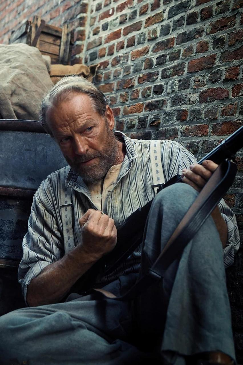 Ein älterer Mann mit Bart hält ein Gewehr. Er sitzt in der Nähe einer Ziegelmauer. Er trägt ein gestreiftes Hemd und Hosenträger.