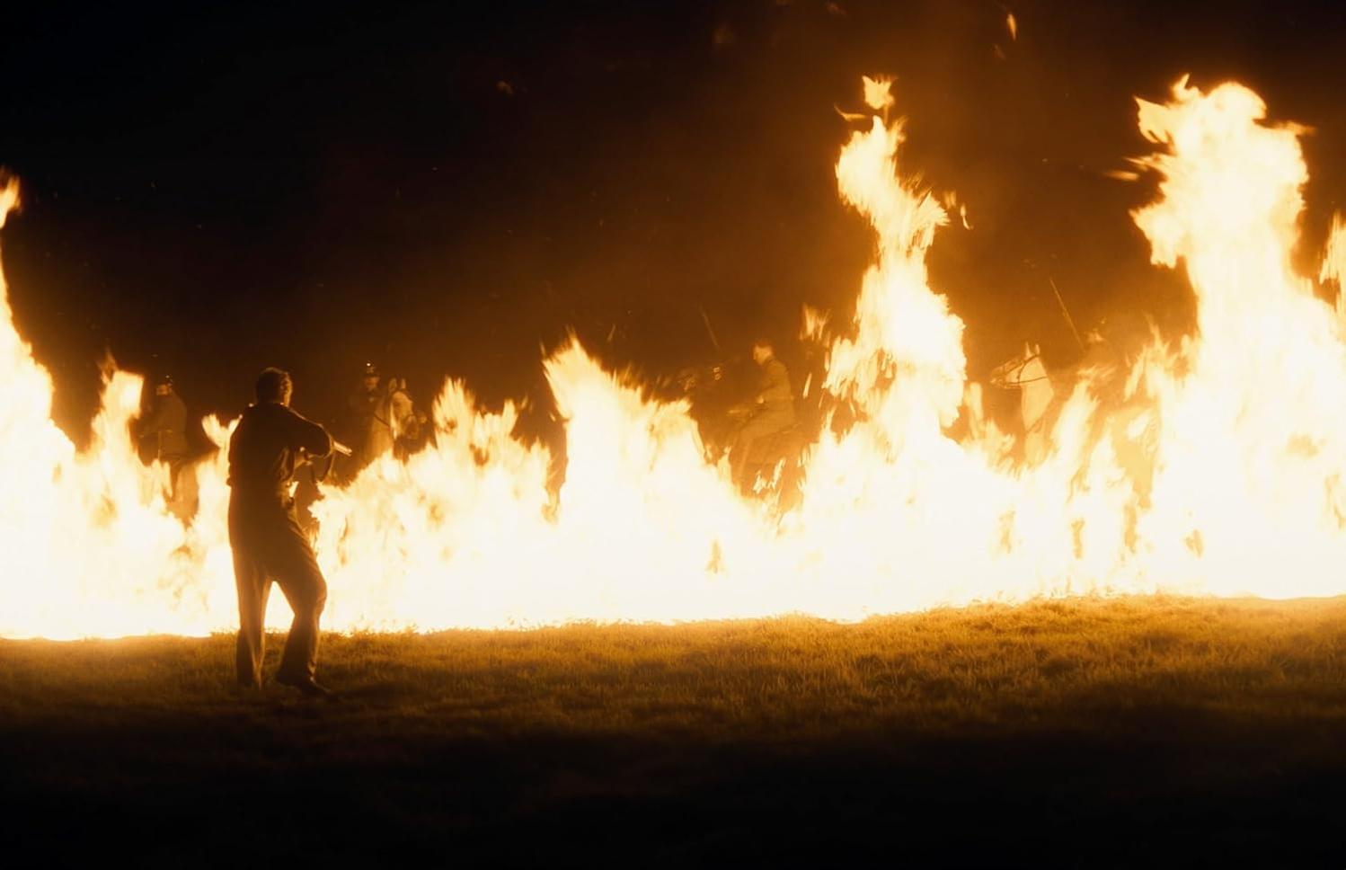Ein Mann steht vor einer Feuerwand, Silhouetten von Menschen in den Flammen. Die Szene ist dunkel, mit einem grasbewachsenen Vordergrund.