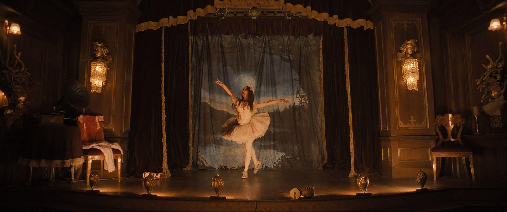 Eine Ballerina tanzt auf einer Bühne. Die Bühne ist mit warmen Lichtern beleuchtet. Der Hintergrund umfasst einen Vorhang und eine bemalte Kulisse.
