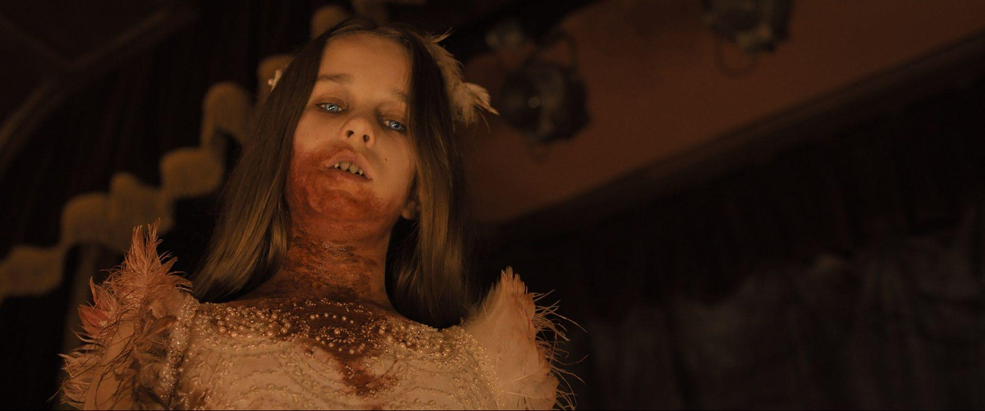 Eine junge Frau mit Blut im Gesicht und auf der Brust, trägt ein weißes Kleid mit Federn und starrt. Dunkler Hintergrund.