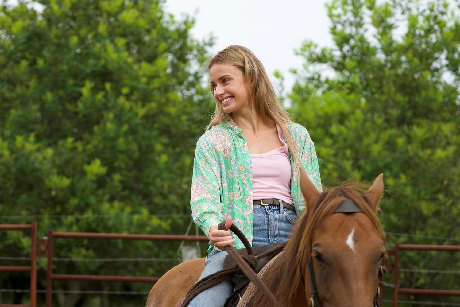 Eine lächelnde Frau reitet auf einem braunen Pferd. Sie trägt ein rosa Top, ein Blumenhemd und Jeans. Im Hintergrund sind grüne Bäume.