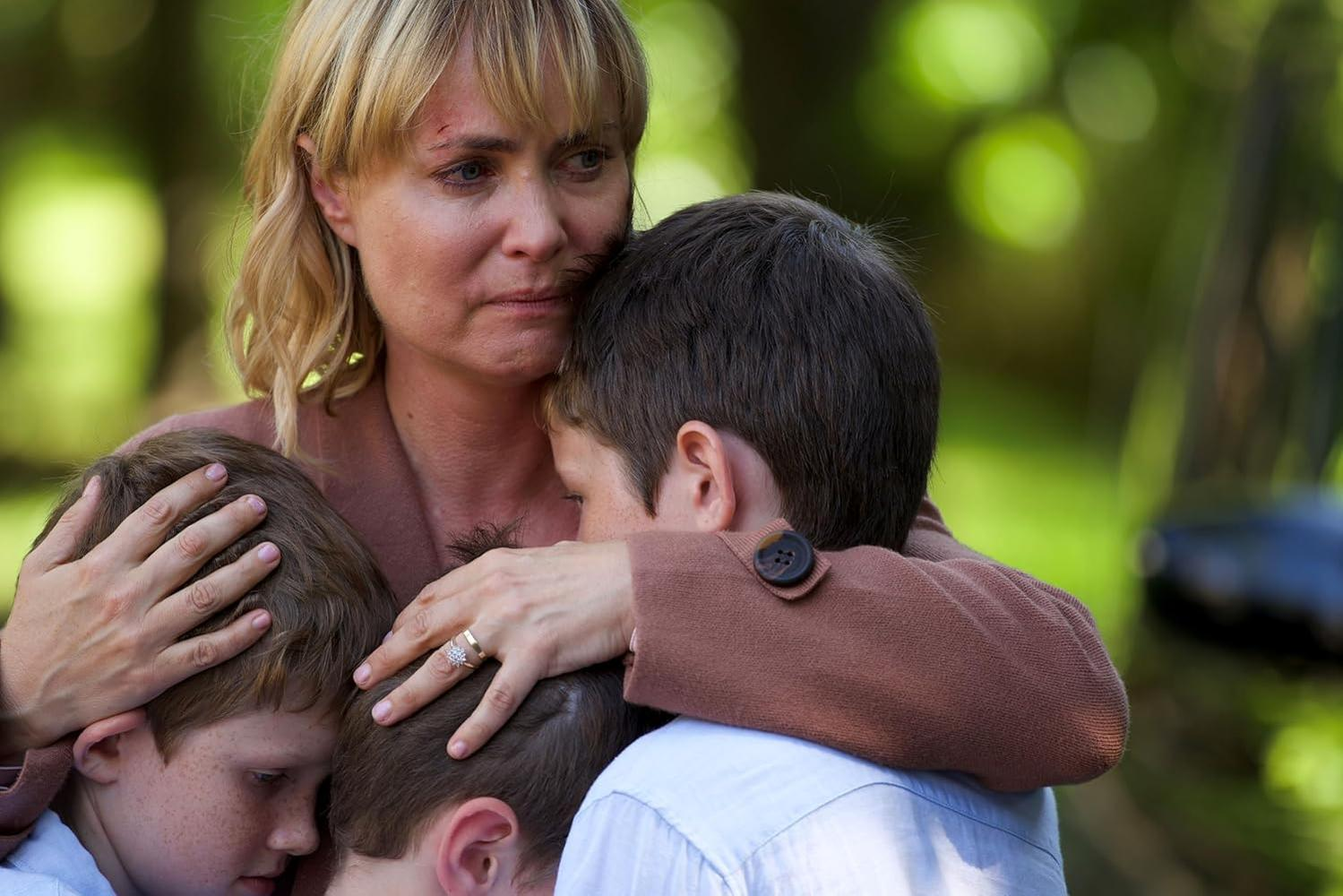 Eine Frau umarmt drei Kinder. Sie hat blondes Haar, einen Bluterguss auf der Stirn und trägt ein braunes Oberteil. Die Kinder sind zusammengedrängt.