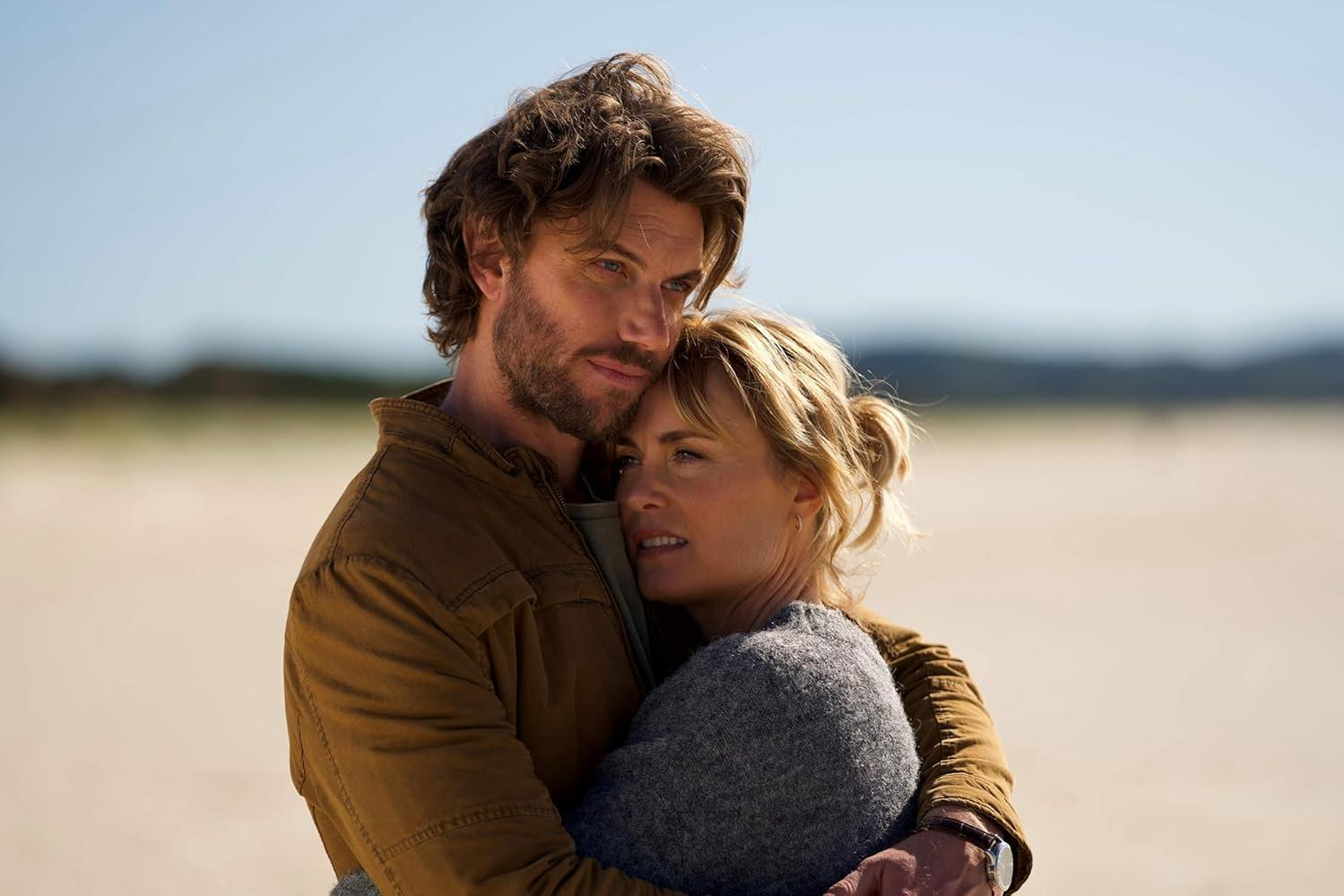 Ein Mann und eine Frau umarmen sich am Strand. Er trägt eine braune Jacke, sie einen grauen Pullover. Klarer blauer Himmel im Hintergrund.