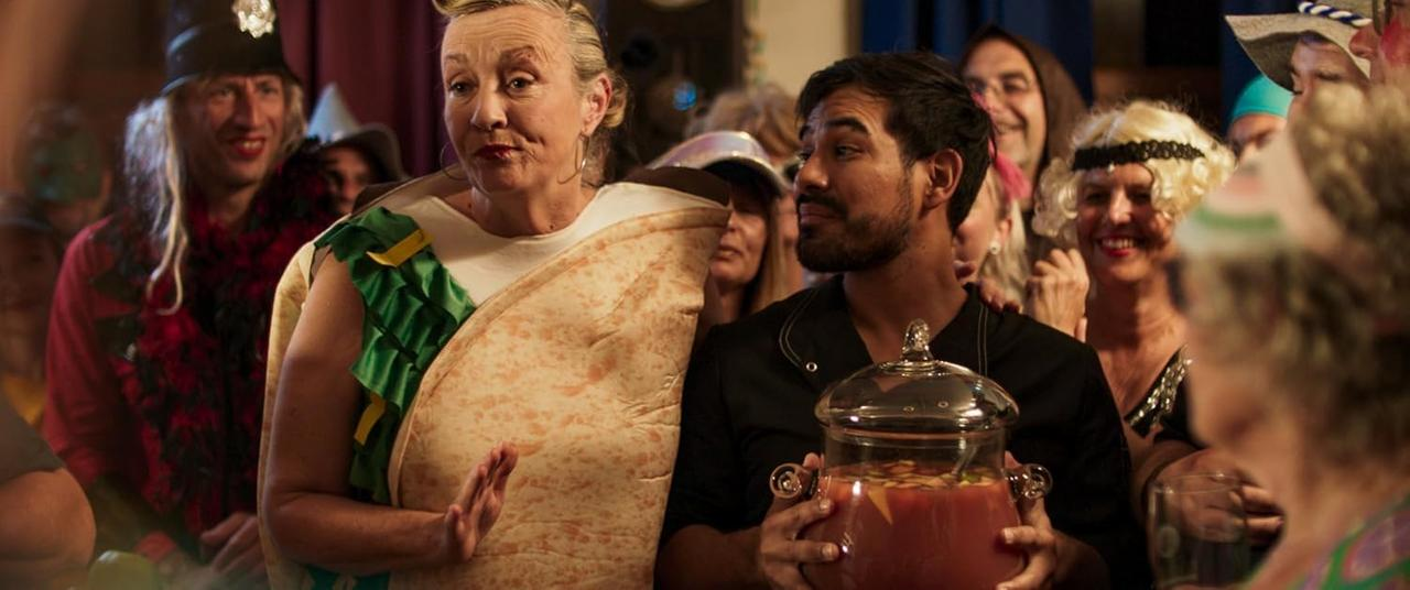 Eine Kostümparty-Szene. Eine Person in einem Taco-Kostüm steht neben einem Mann, der eine Bowle hält. Andere verkleidete Personen sind im Hintergrund.