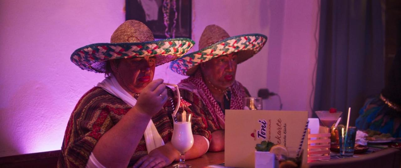 Zwei Personen mit Sombreros an einer Bar, eine hält ein Glas weißen Cocktails. Eine Speisekarte ist sichtbar, und es gibt andere Getränke.