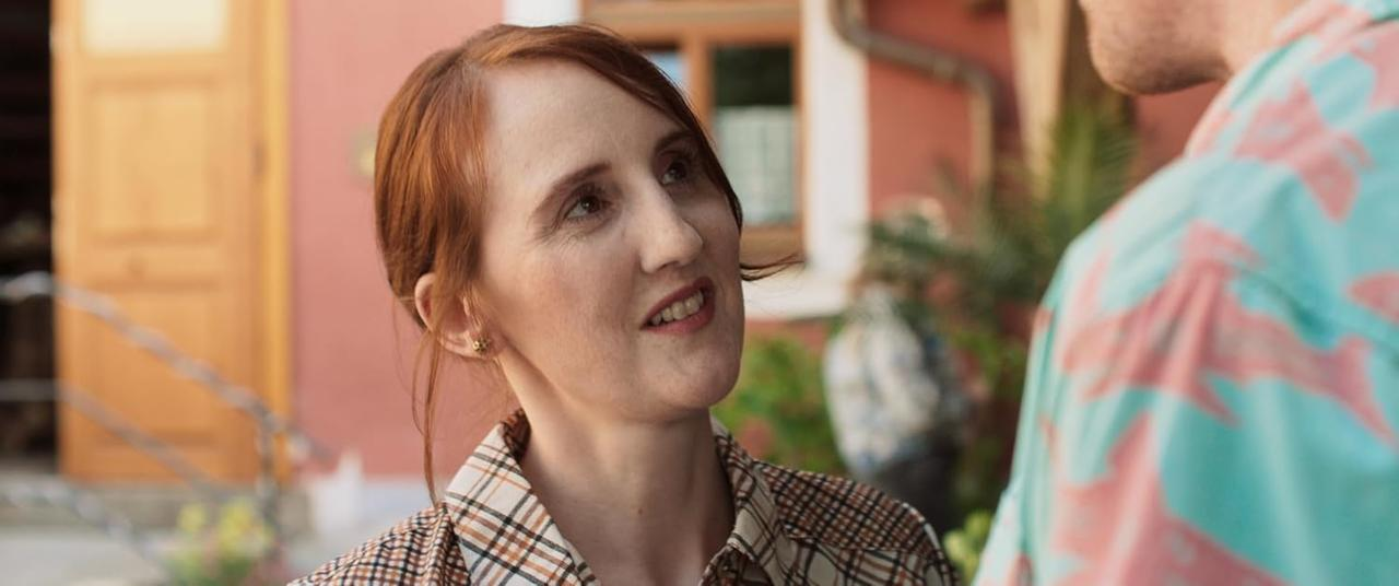 Eine rothaarige Frau mit kariertem Hemd spricht mit jemandem. Ein Mann in einem Hemd mit Blumenmuster ist teilweise sichtbar.