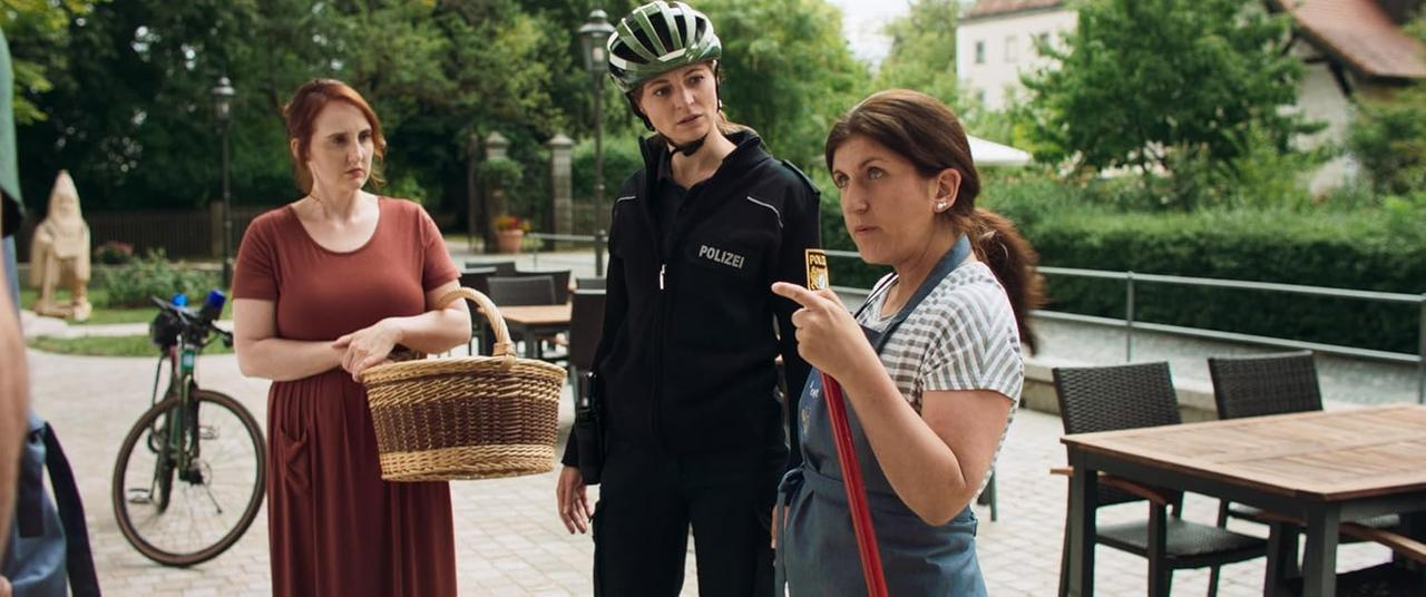 Drei Frauen draußen: eine mit Korb, eine in Polizeiuniform und eine zeigt. Ein Fahrrad ist ebenfalls vorhanden.