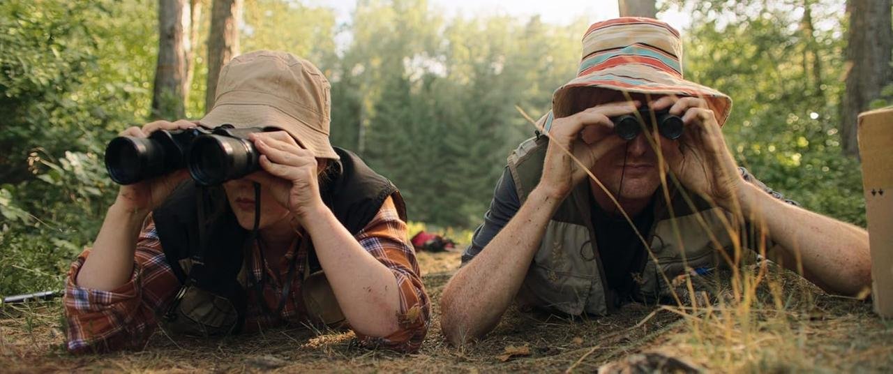 Zwei Personen liegen auf dem Boden und schauen durch ein Fernglas. Sie tragen Hüte und befinden sich in einem Wald.