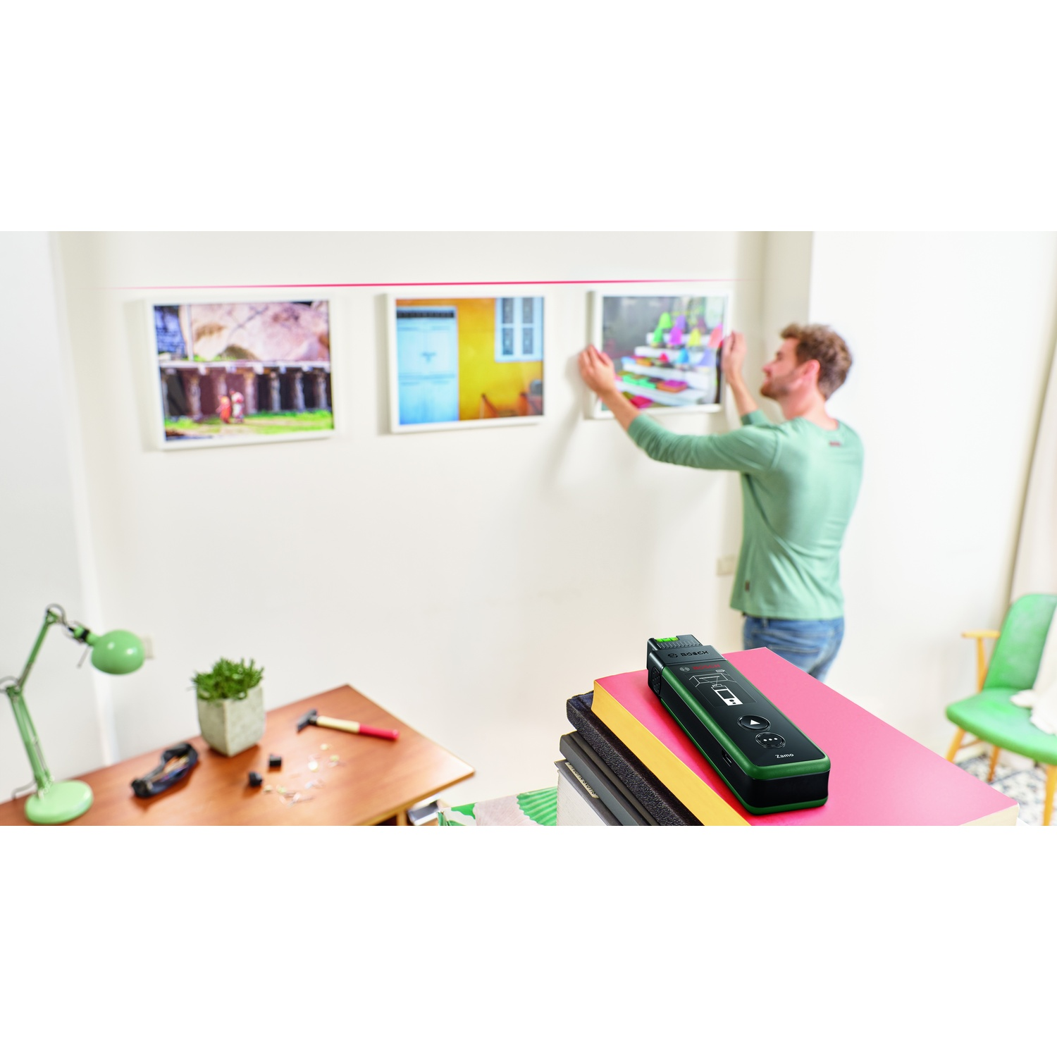 Un uomo appende un quadro al muro, ci sono già due quadri appesi. Attrezzi e libri in primo piano.