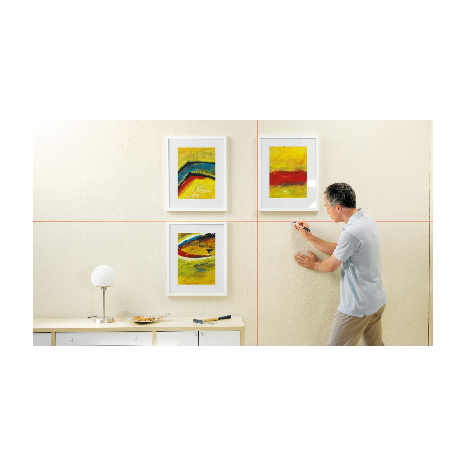 Un uomo sta tracciando una linea su un muro beige. Tre opere d'arte astratte incorniciate sono appese al muro. Una lampada e un martello sono su un mobile.