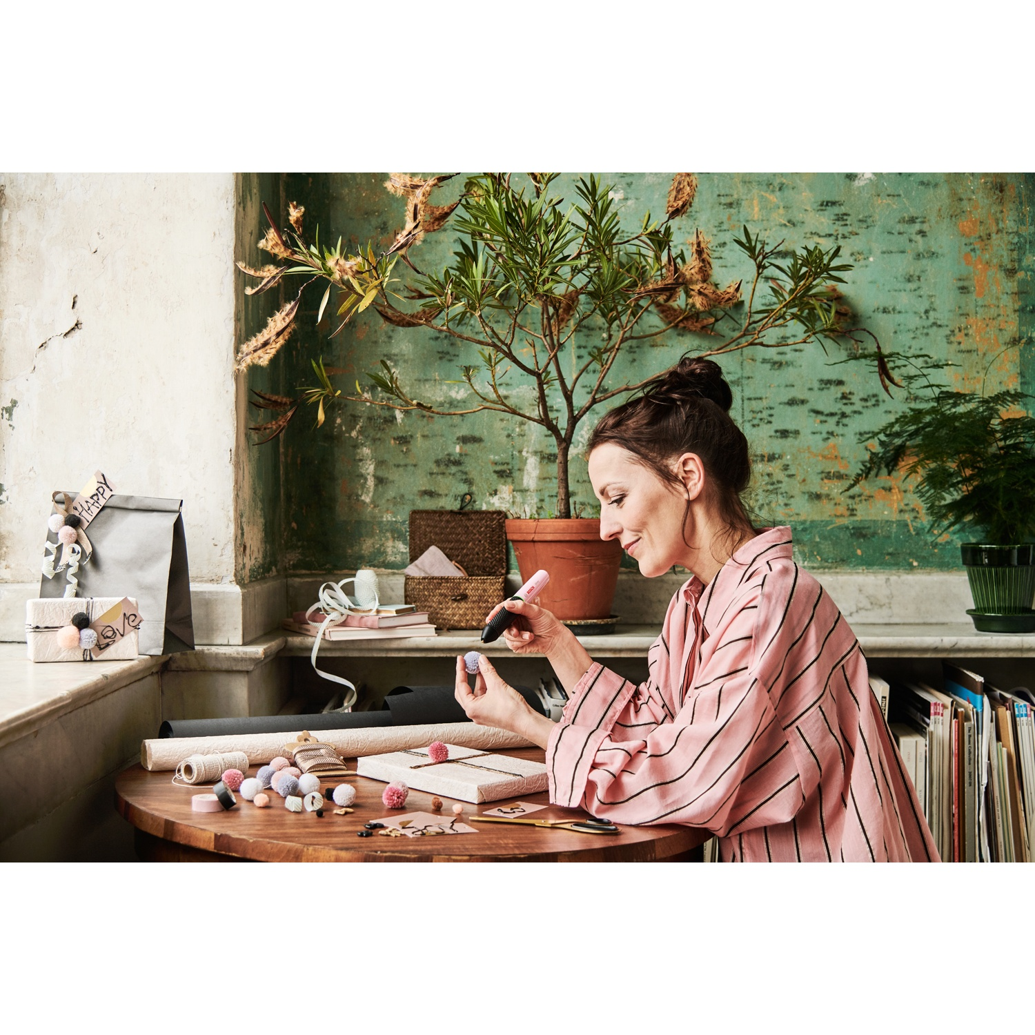 Donna con camicia rosa a righe che crea su un tavolo di legno. Ci sono forniture per l'artigianato, una pianta e una finestra sullo sfondo.