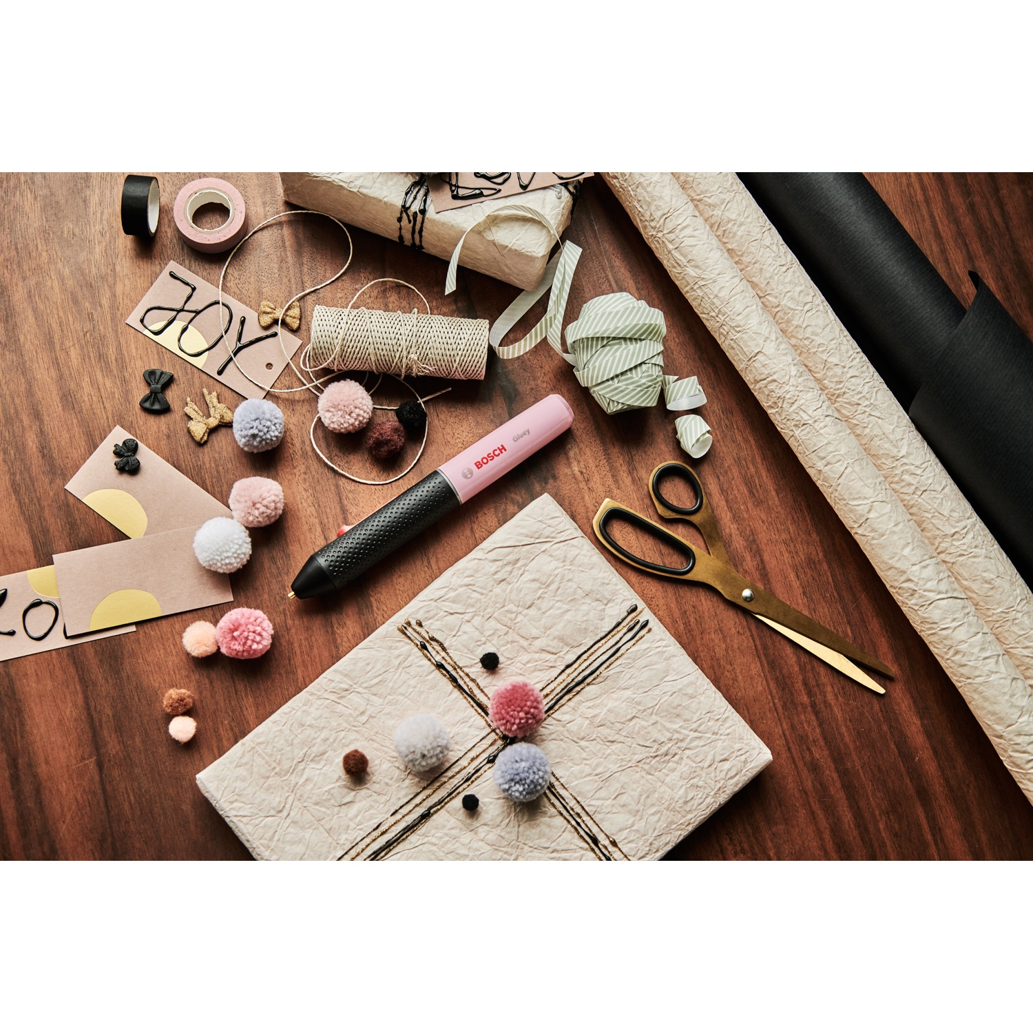 Una superficie in legno con materiali per confezioni regalo. Include una pistola per colla, carta da regalo, forbici, pompon, nastro adesivo e confezioni regalo.