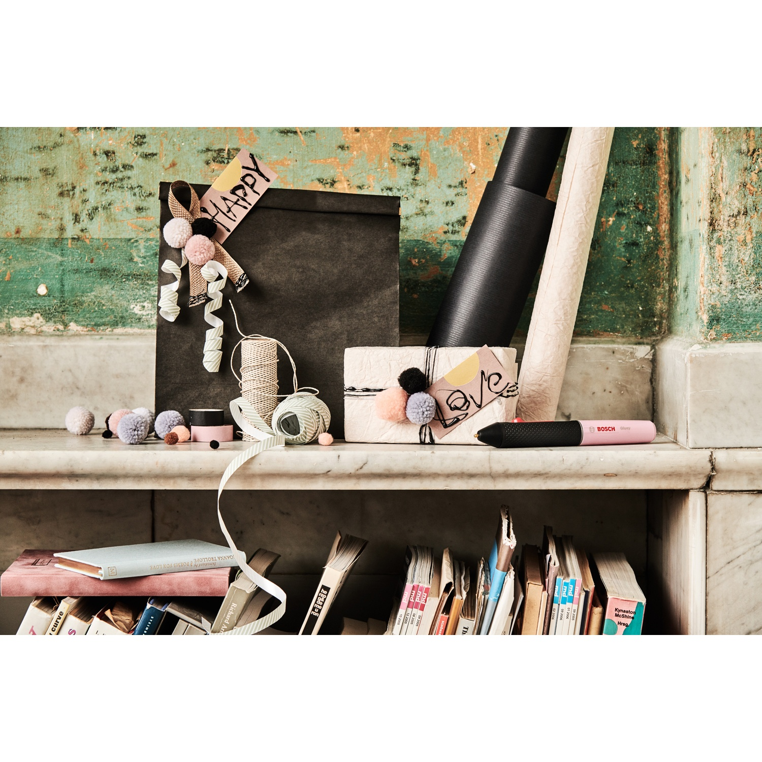 Scaffale con materiali per confezioni regalo, libri e una penna colla rosa BOSCH. Sono presenti anche elementi decorativi come pompon e nastri.
