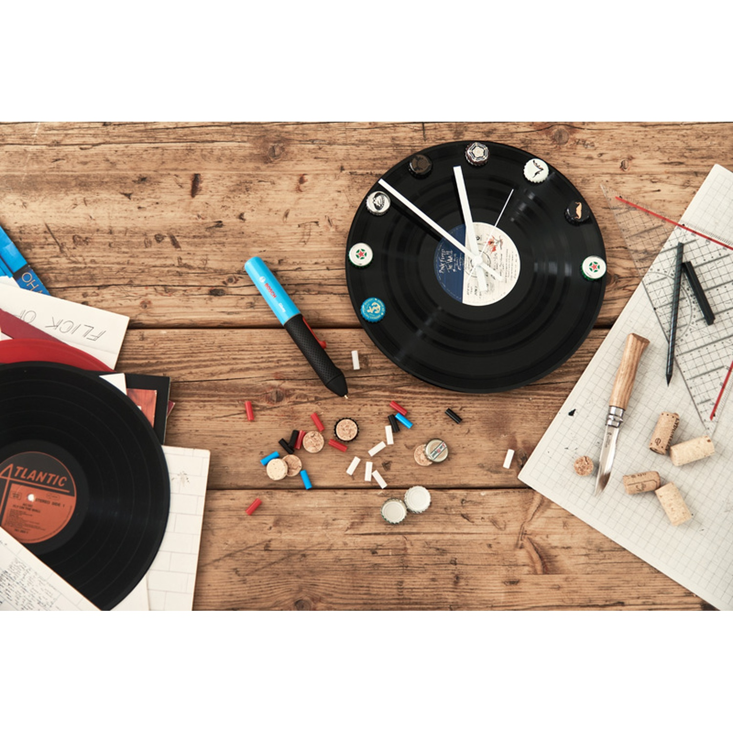 Un orologio in vinile con tappi di bottiglia come numeri. Un disco, una penna, dei tappi e un righello su un tavolo di legno.